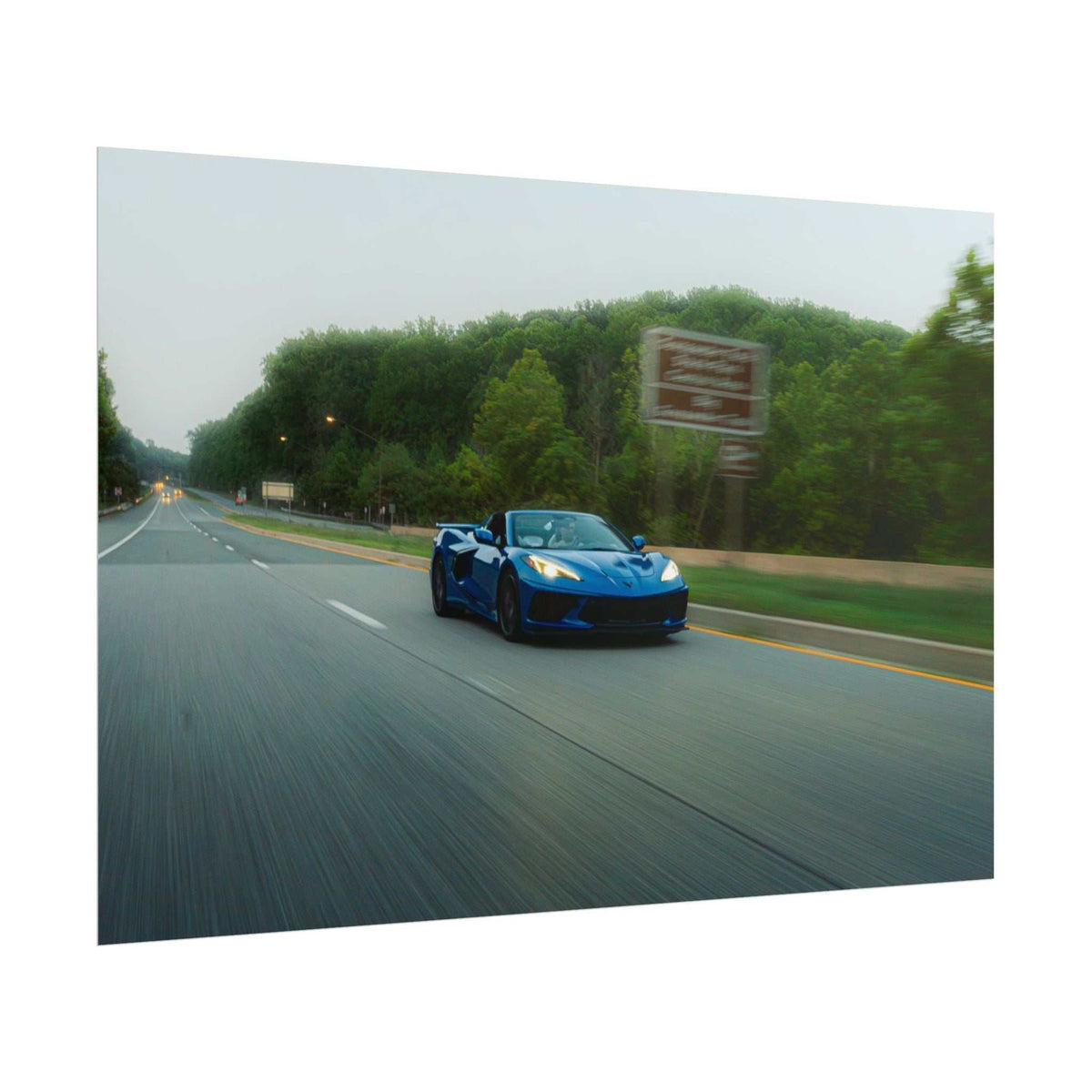 C8 Corvette Stingray in blue driving on a multi-lane highway surrounded by green trees under a cloudy sky