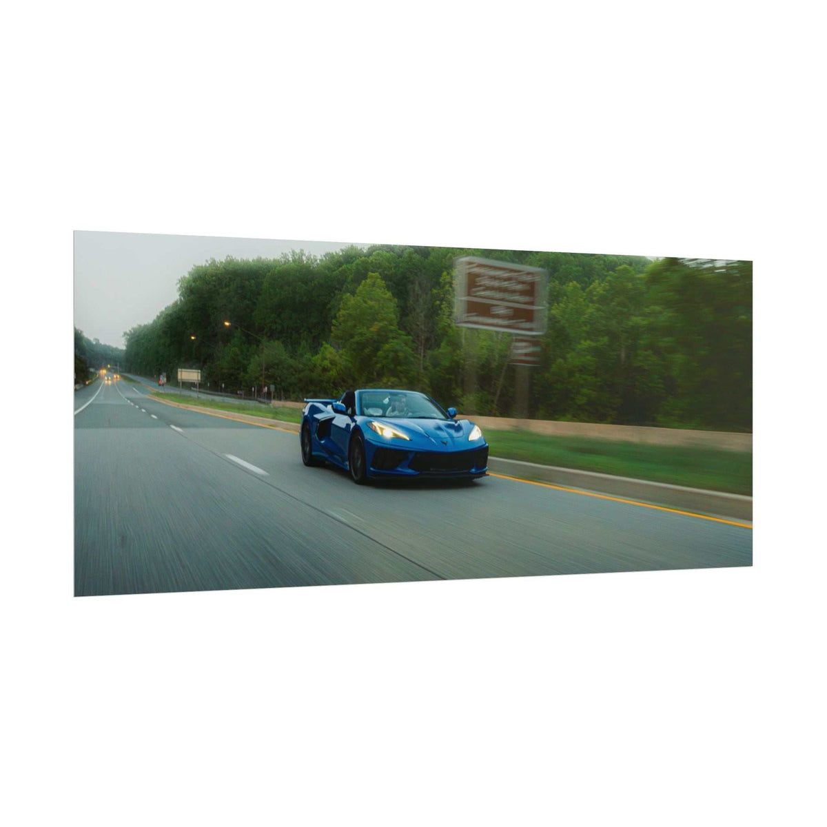 Blue C8 Corvette Stingray sports car driving on a tree-lined highway with motion blur capturing speed and depth