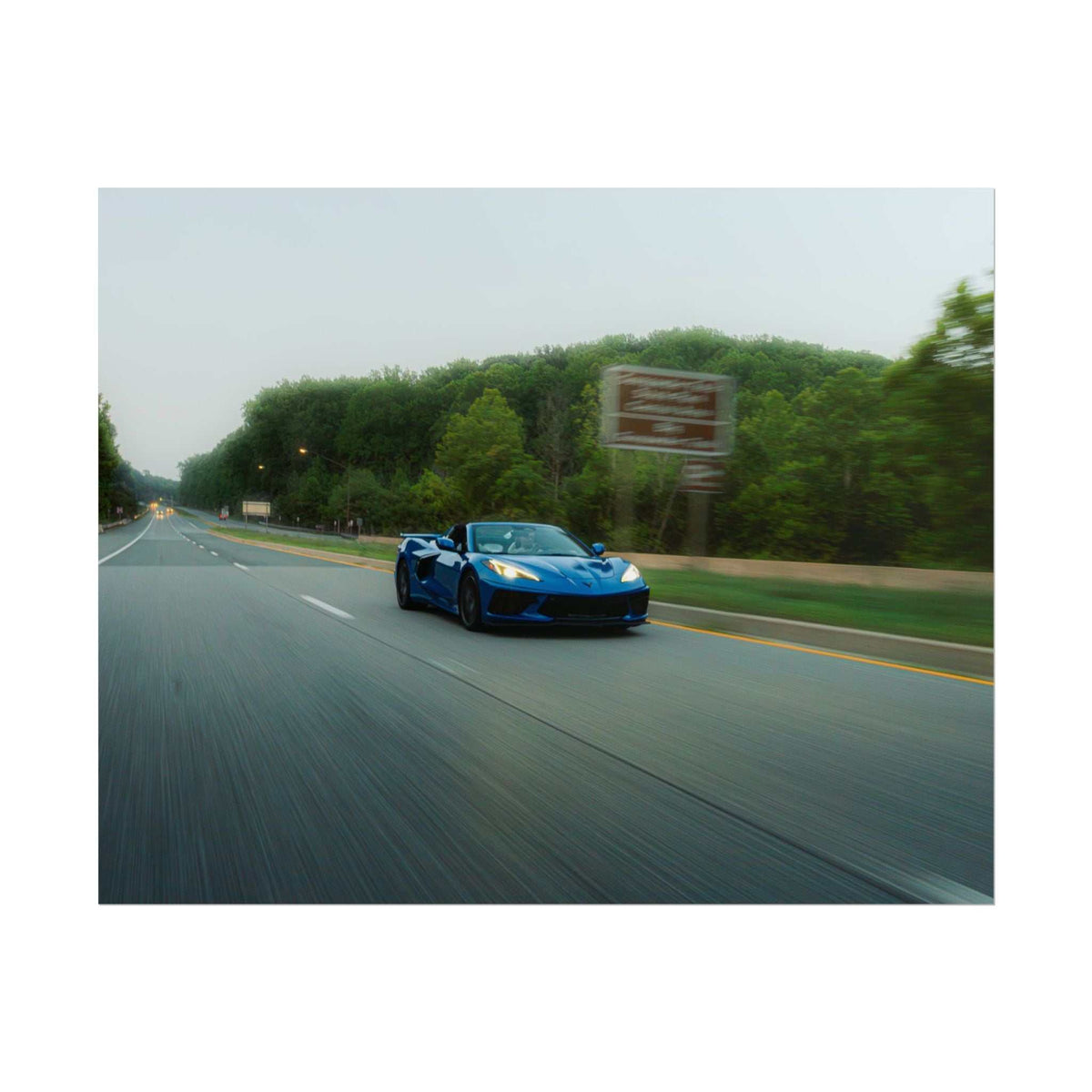 Blue C8 Corvette Stingray sports car driving on a highway surrounded by green trees with motion blur effect