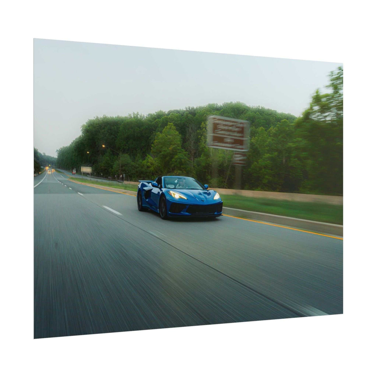 C8 Corvette Stingray sports car in blue captured in motion on a tree-lined highway with speed blur and natural surroundings
