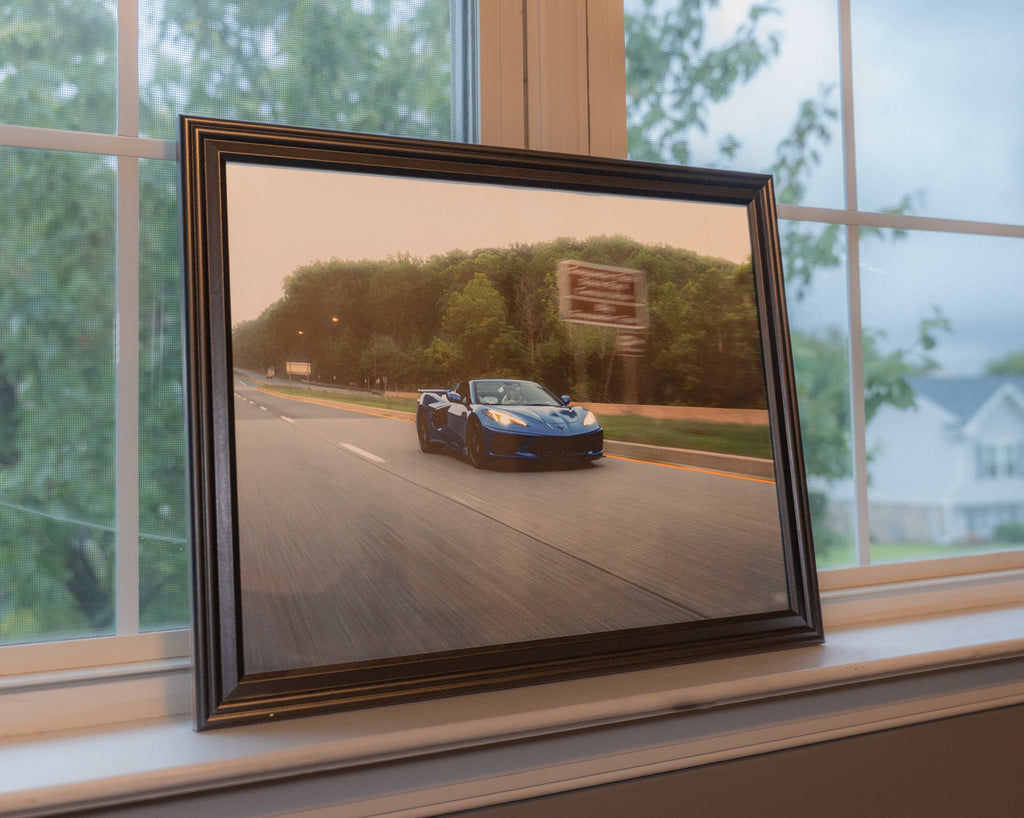 Framed art print of a blue C8 Corvette Stingray sports car driving on a blurred highway with green trees background