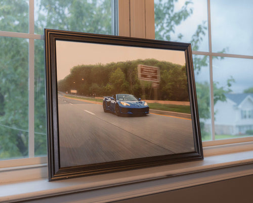 Framed art print of a blue C8 Corvette Stingray sports car driving on a blurred highway with green trees background